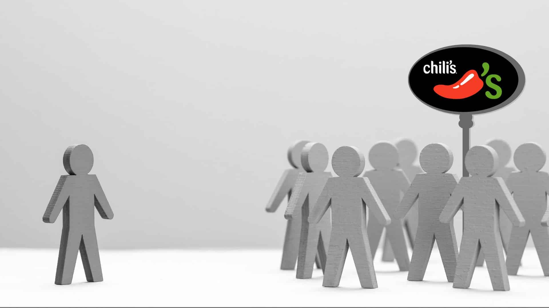 wooden people depicting discrimination by showing one to the left and a group of others to the right, a Chili's sign is behind the group