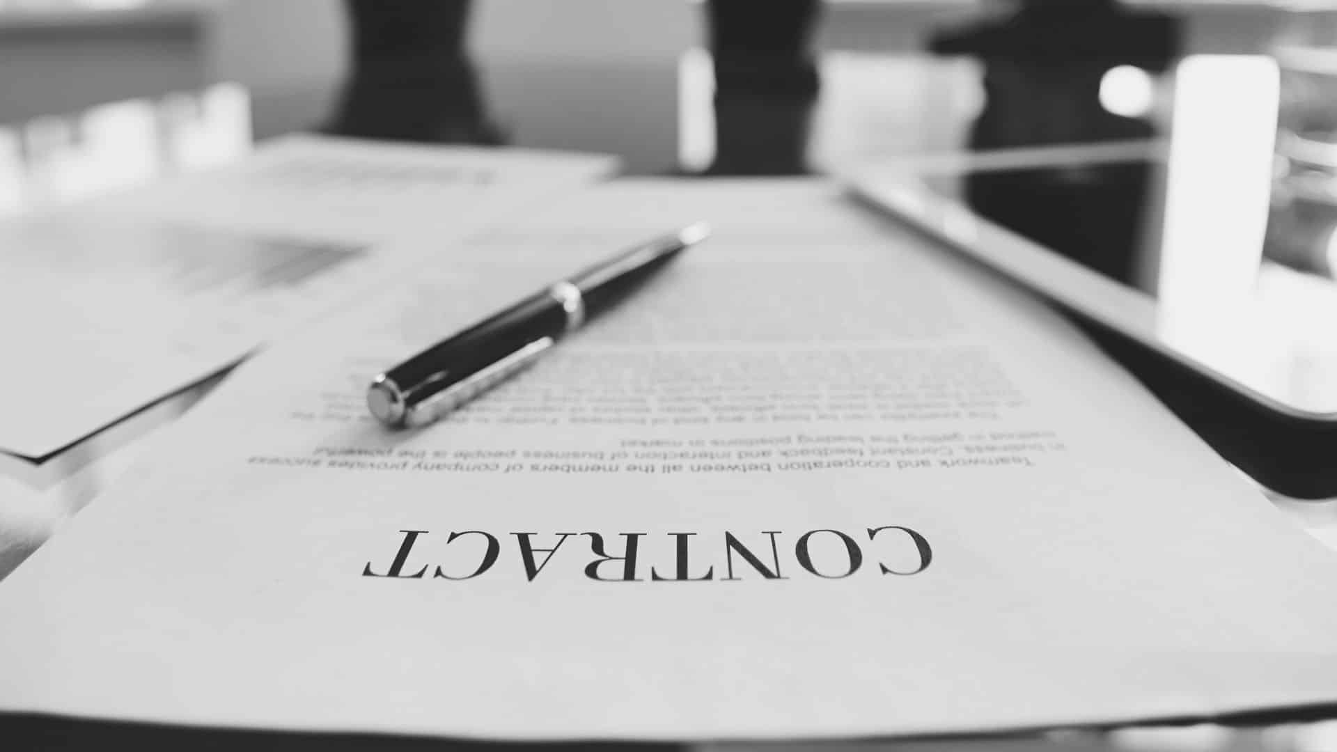 paper sitting on a table, facing away from the camera, reading "contract" with a pen sitting on the paper