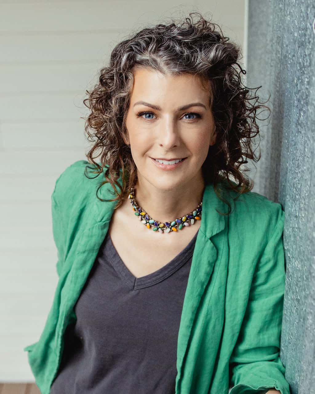 Woman with curly hair leaning against a wall, looking up at the camera while wearing a green blazer and dark gray v-neck