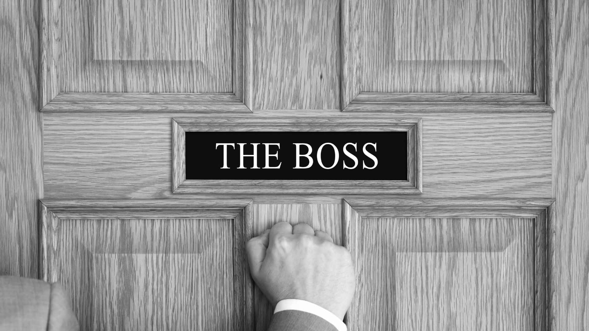 hand knocking on a door, on the door is a small sign that reads "THE BOSS"